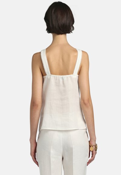 Femme aux cheveux courts et foncés portant un débardeur blanc à larges bretelles et un pantalon blanc, avec des bracelets dorés au poignet droit.