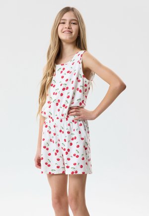 Tienermeisje met lang haar, gekleed in een wit mouwloos topje en korte broek met rood kersenpatroon, staand met één hand op haar heup.