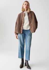 Bruine fleecejas met een satijnen voering en drukknoopsluitingen, gedragen over een crèmekleurige trui, gecombineerd met blauwe straight-leg jeans en zwarte enkellaarsjes.