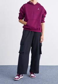 Sudadera burdeos con bolsillo frontal y logo, combinada con pantalones cargo oscuros que tienen bolsillos laterales. Usada con zapatillas burdeos y blancas.