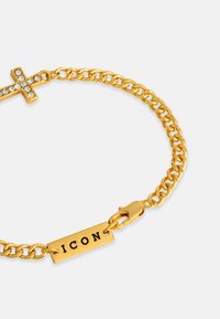 Bratara din aur cu un crucifix accentuat de diamante și o etichetă rectangulară gravată cu "ICON". Textură metalică netedă.