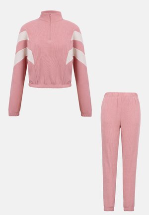Survêtement deux pièces rose avec des rayures en chevron blanches sur les manches, comprenant un haut zippé court à manches longues et un pantalon jogging taille élastique.