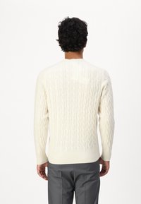 Cream cable-knit sweater with ribbed cuffs and hem. Worn with gray trousers, showcasing a textured pattern on the fabric.