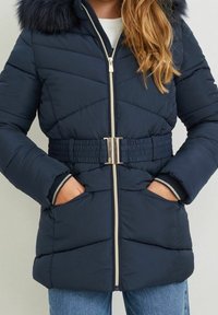 Chaqueta de plumón azul marino con capucha forrada de piel, cintura ajustada con cinturón, cierre de cremallera y puños ribeteados. Cuenta con un patrón de acolchado diagonal y dos bolsillos.