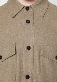 Tan wollen overhemd met een kraag, voorzien van zwarte knopen en twee borstzakken. De stof heeft een zachte textuur en een gladde afwerking.