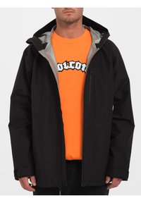 Czarna kurtka z zamkiem z przodu, kapturem i kontrastowym szarym wykończeniem, noszona na pomarańczowym t-shircie z grafiką. Gładka faktura, casualowy design.