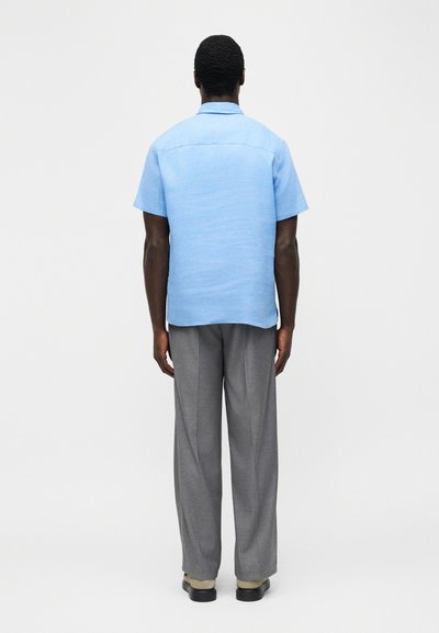 Άνδρας με την πλάτη, φοράει ανοιχτό γαλάζιο κοντομάνικο πουκάμισο, γκρι παντελόνι και μαύρα παπούτσια, πάνω σε απλό ανοιχτό φόντο.