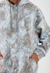 Sivé a hnedé nadmerné mikiny s abstraktným mramorovým vzorom, so predným vreckom a kapucňou na šnúrku. Mäkkej, textúrovanej látky.