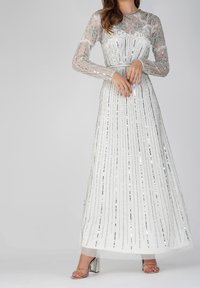 Lungo vestito bianco con maniche lunghe in voile, perline argentate e paillettes a strisce verticali. Presenta un corpetto aderente e una gonna fluida. Tacchi argentati.