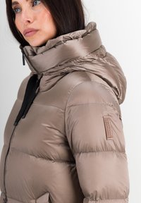 Zacht beige jasje met een hoge kraag en capuchon, voorzien van een zwarte rits en logo-tag op de mouw. Glad, glanzend oppervlak.
