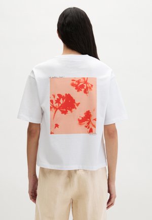 Femme aux longs cheveux foncés portant un t-shirt blanc avec une grande impression florale rouge dans le dos et un pantalon beige, debout devant un fond uni.