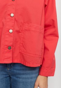 Röd knapplistjacka i lätt tyg med en stor framficka, synlig sömnad och en avslappnad passform.