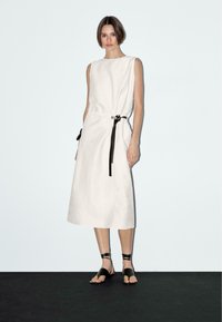 Beige linen dress with a loose fit, sleeveless design, and a black tie at the waist. Features flat black sandals. Simple, minimalist style.