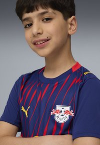 Sinine ja punane triibuline Puma spordijerseys, millel on ümar kaelus, RB Leipzigi logo ja kollased aktsendid õlgadel.