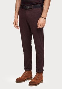 Bordowe spodnie z teksturowanej tkaniny, o zwężanym kroju i podwiniętych nogawkach, zestawione z brązowymi zamszowymi butami na sznurowadła oraz czarnym paskiem.