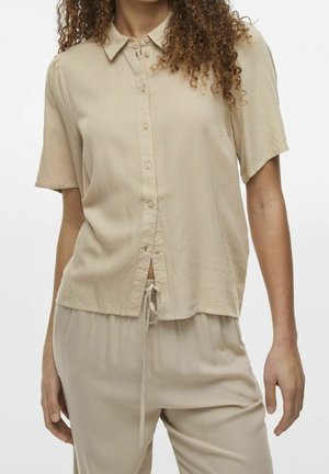 Vrouw die een beige kortemouwen blouse met knopen draagt en bijpassende broek met koord, weergegeven van schouders tot halverwege het dijbeen.