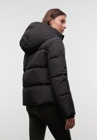 Frau in schwarzer, gepolsterter Kapuzenjacke und dunkler Leggings, seitlich vor einem schlichten, hellen Hintergrund stehend.