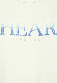White sweatshirt featuring "HEAR THE SEA" in gradient blue sequins with a subtle sheen. Soft texture and casual design.