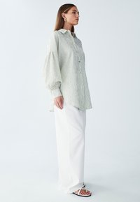 Prevelika srajca z geometrijskimi trakovi in napihnjenimi rokavi, svetlo zelene in bele barve, kombinirana z belimi širokimi hlačami in ravnimi sandali.