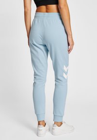 Lyseblå joggingbukser lavet af blødt stof, med et hvidt chevronmønster på venstre ben og ribbede manchetter. Sammen med hvide sneakers.