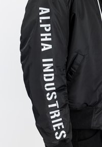Zwarte jas mouw met witte verticale "ALPHA INDUSTRIES" tekst langs de arm gedrukt, met geribde manchetten en zijzak met drukknop.