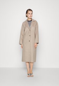 Beige wolcoats met een notch-kraag en drie knopen, met een relaxed fit en lange mouwen, gelaagd over een zwart-wit gestreepte top.
