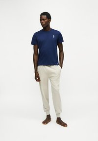 Homme debout pieds nus portant un t-shirt bleu foncé avec un petit logo et un pantalon de survêtement gris clair sur fond blanc uni.