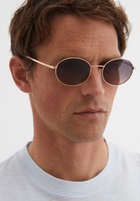 Lunettes de soleil avec des verres ronds et sombres, fines montures en métal rose gold, et branches minimalistes. La monture est élégante avec un branding subtil.