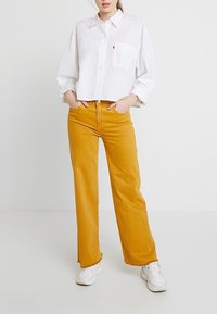 Vit kortärmad skjorta med knappar och bröstficka, tillsammans med högmidjade vida jeans i senapsgul färg. Modellen har på sig vita sneakers.