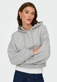 Femme aux cheveux châtain clair portant des boucles d'oreilles en argent, un sweat à capuche gris, une chemise blanche et un jean bleu marine, les bras croisés.