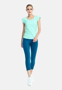 Mintgroene top met korte mouwen en een ronde hals, gecombineerd met teal geknipte leggings. De outfit wordt compleet gemaakt met witte sportschoenen.