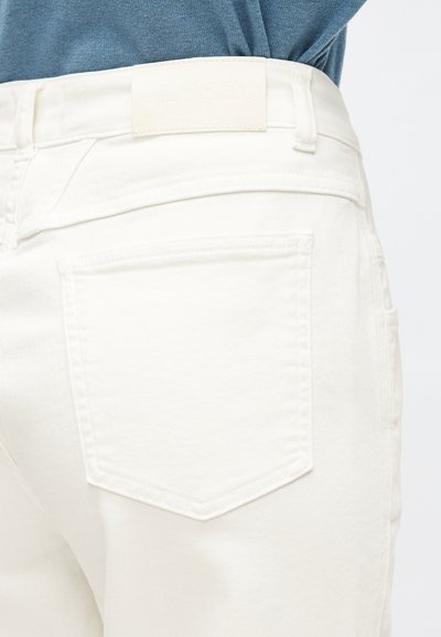Gros plan sur un pantalon en denim blanc avec une poche arrière et des passants de ceinture, porté avec une chemise bleue rentrée.