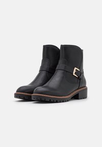 Botas de piel negras hasta el tobillo con un tacón medio tipo bloque, con una correa lateral y un hebilla en tono dorado, suela texturizada y diseño de punta redonda.