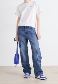 Hvid bomulds t-shirt med lille cirkellogo, blå denim cargo bukser med flere lommer, lilla crossbody taske og blå og hvide sneakers.