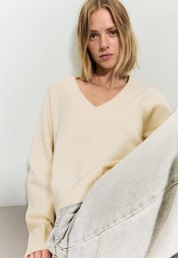 Mlada ženska s srednje dolgimi blond lasmi, oblečena v kremno V-izrez pulover in svetlo sivo jeans, sedi ob navadni beli steni.
