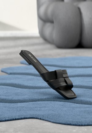 Sandalo nero slip-on con cinturini intrecciati che poggia su un tappeto blu testurizzato, con un oggetto grigio arrotondato sfocato sullo sfondo.