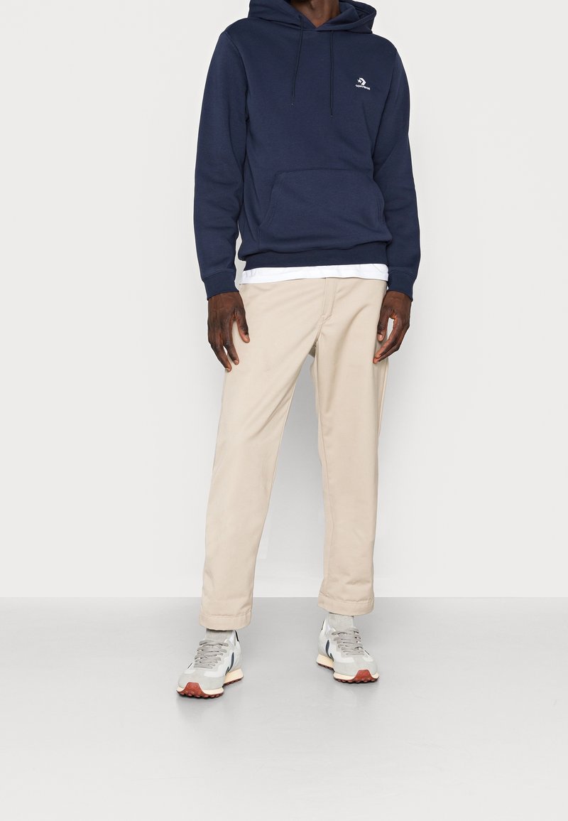 Personne debout devant un fond uni, portant un sweat à capuche bleu marine, un pantalon beige et des baskets gris-blanc à semelles orange. Le haut du visage n'est pas visible.