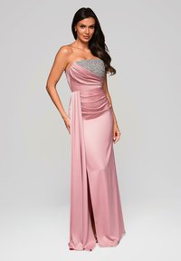 Kvinna i en strapless rosa satängklänning med en silverpärlad överdel, stående med en hand på höften mot en enkel ljus bakgrund.