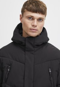 Jachetă puffer neagră cu guler înalt, având două buzunare laterale cu fermoar, design matlasat și o textură netedă, mată.