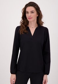 Vrouw met golvend bruin haar, die een zwarte blouse met lange mouwen en een V-hals draagt, en een fijn kettinkje om haar hals, staand tegen een effen achtergrond.