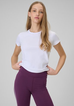 Młoda kobieta o długich włosach, ubrana w dopasowany biały t-shirt z krótkim rękawem i fioletowe legginsy, stoi z rękami na biodrach na szarym tle.