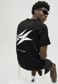 Sort atletisk t-shirt med korte ærmer, der har et stort hvidt grafisk design på ryggen. Et par sorte shorts og solbriller fuldender looket.