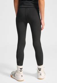 Schwarze Leggings mit glatter Textur, figurbetontem Schnitt und kontrastierendem Chevron-Muster an der Seite. Kombiniert mit weißen Sportschuhen und Socken.