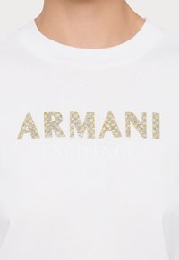 Vit bomulls-T-shirt med "ARMANI" i guld- och silverstrass och "EXCHANGE" i subtil vit text nedanför. Kortärmad och rund hals.