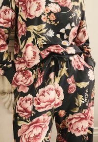 Souprava domácího oblečení s květinovým vzorem v černé barvě, která má velké růžové pivoňky a akcenty zelených listů. Obsahuje pásky na stažení v pase a na manžety.