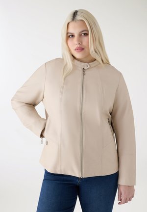 Kobieta z długimi blond włosami, ubrana w beżową kurtkę na zamek i niebieskie jeansy, stoi z jedną ręką na biodrze na jednolitym tle.