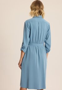Jasnoniebieska sukienka koszulowa z kołnierzykiem, zapięciem na guziki przy mankietach i paskiem w talii. Charakteryzuje się luźnym krojem i średnią długością z bocznymi rozcięciami.