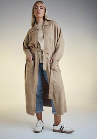 Ljusbrun stickad cardigan med bälte, stora fickor, ribbade ärmar, bärs över en beige topp och i kombination med blå cropped jeans och sneakers.