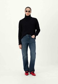 Mulher com cabelo trançado vestindo um suéter preto de gola alta, jeans azuis, sapatos vermelhos e óculos de sol pretos pequenos, de pé com uma das mãos no bolso.