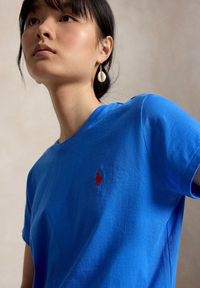 Junge Frau mit dunklen Haaren trägt ein hellblaues T-Shirt mit einem kleinen roten bestickten Polospieler und einem Muschel-Ohrring.